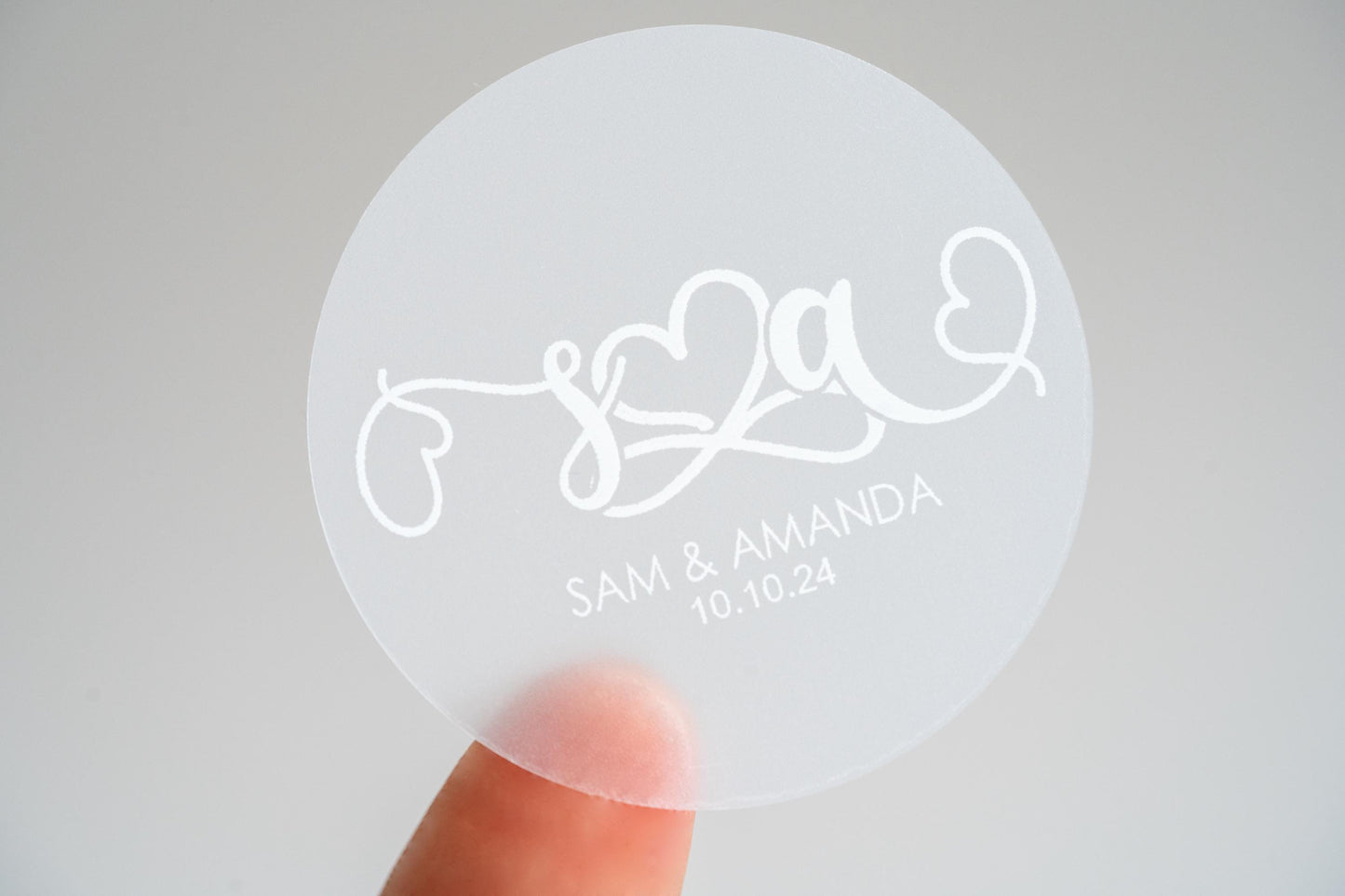 This elegant frosted clear monogram sticker features personalised initials in a stylish handwriting font, crafted in white. An infinity symbol at the centre signifies eternal love, flanked by charming hearts. Ideal for custom gifts or wedding favours