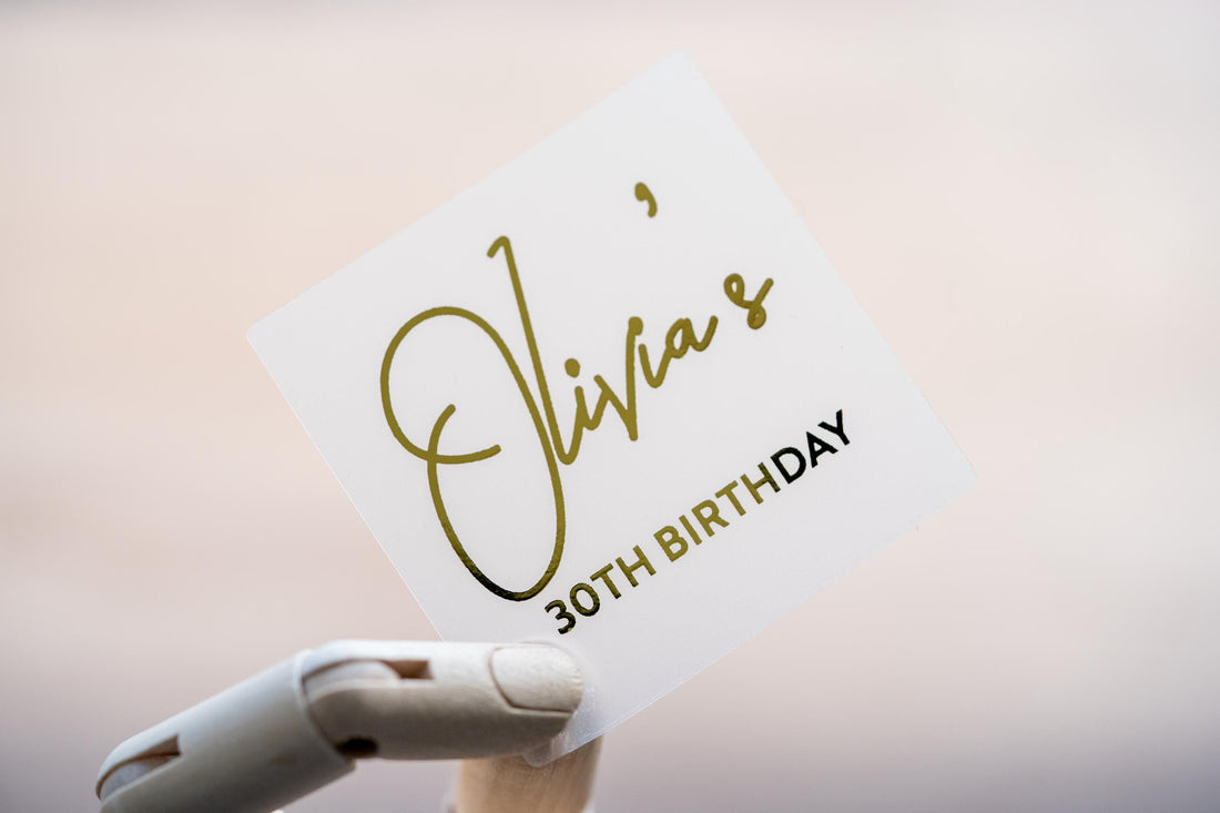 A square sticker with the birthday person's name in a lovely handwritten font. The age is written under the name in a straight font and the entire design layered in a lovely gold foil. The foil shines perfectly in light.
