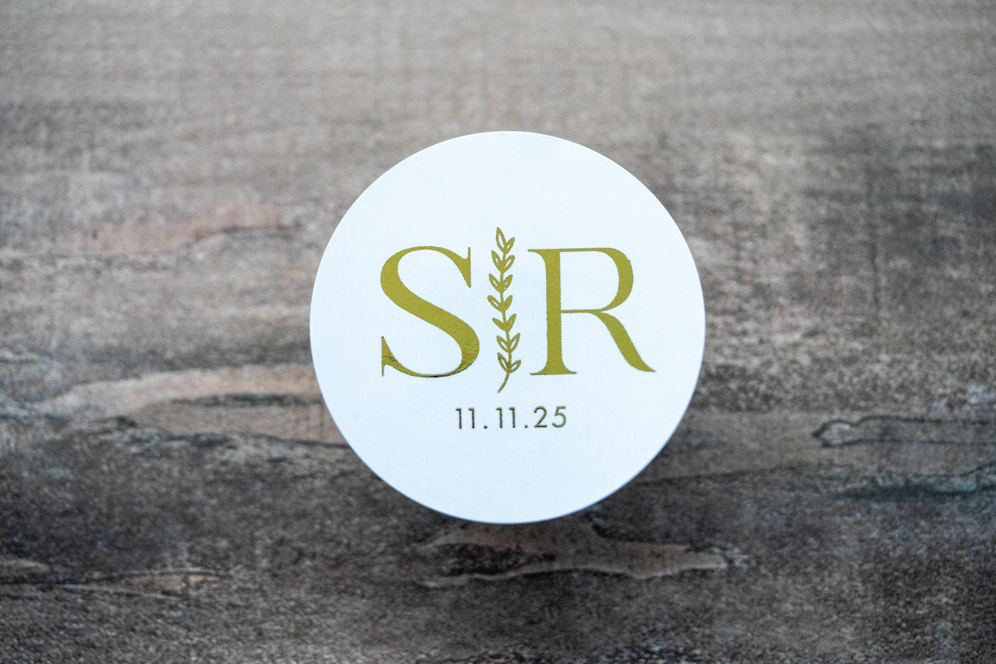A custom round monogram sticker with a leaf border line running vertically across the middle. The initials of the bride and groom sit either side of the leaf border line with the date at the bottom. The design is finished with a lovely metallic foil.