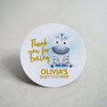 A baby shower sticker with a lovely colour print of an zebra sitting on the floor. Thank you for coming is written in a handwriting font next to the zebra and the mother to be name on the bottom. The writing is finished with metallic foil