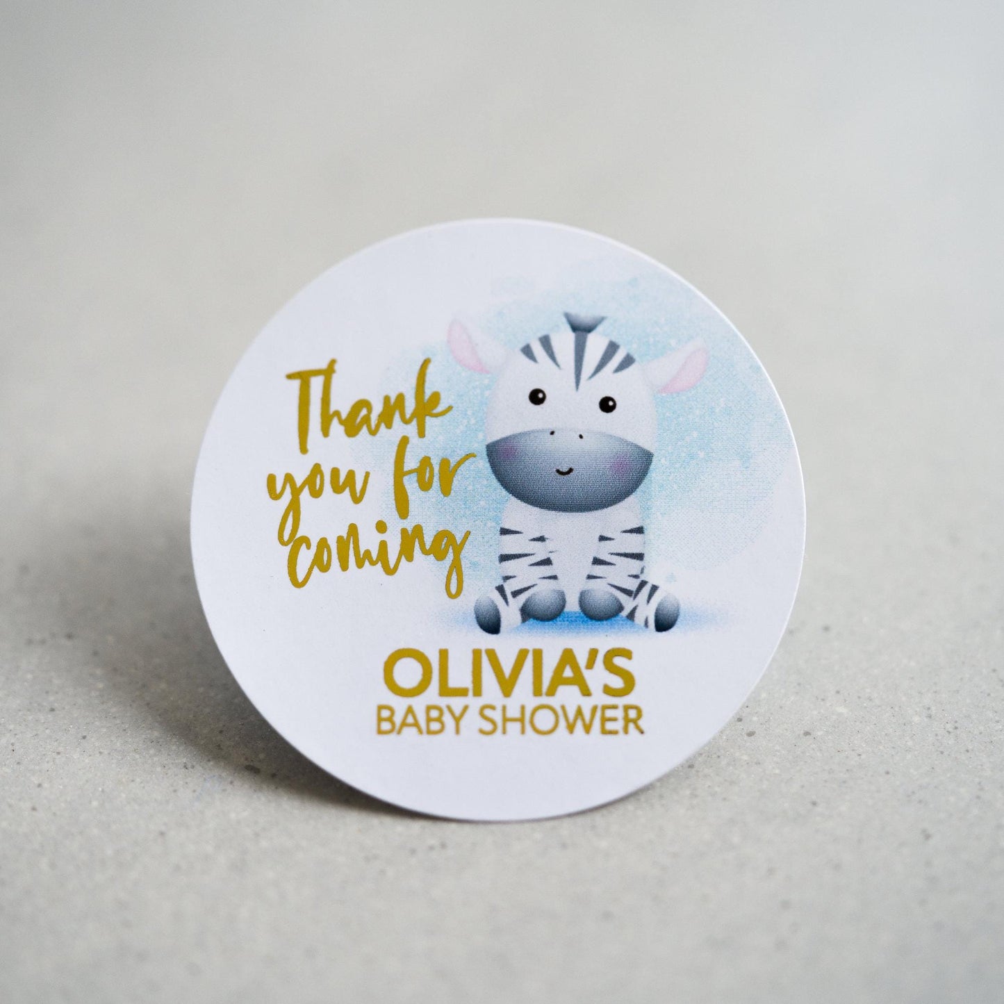 A baby shower sticker with a lovely colour print of an zebra sitting on the floor. Thank you for coming is written in a handwriting font next to the zebra and the mother to be name on the bottom. The writing is finished with metallic foil