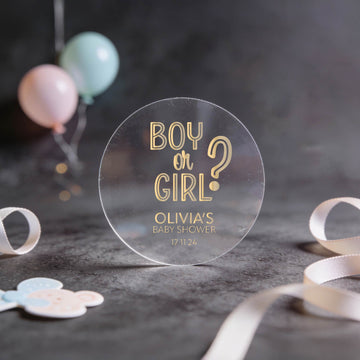 Transparent round sticker with 'Boy or Girl?' question and baby shower details written in gold foil. 