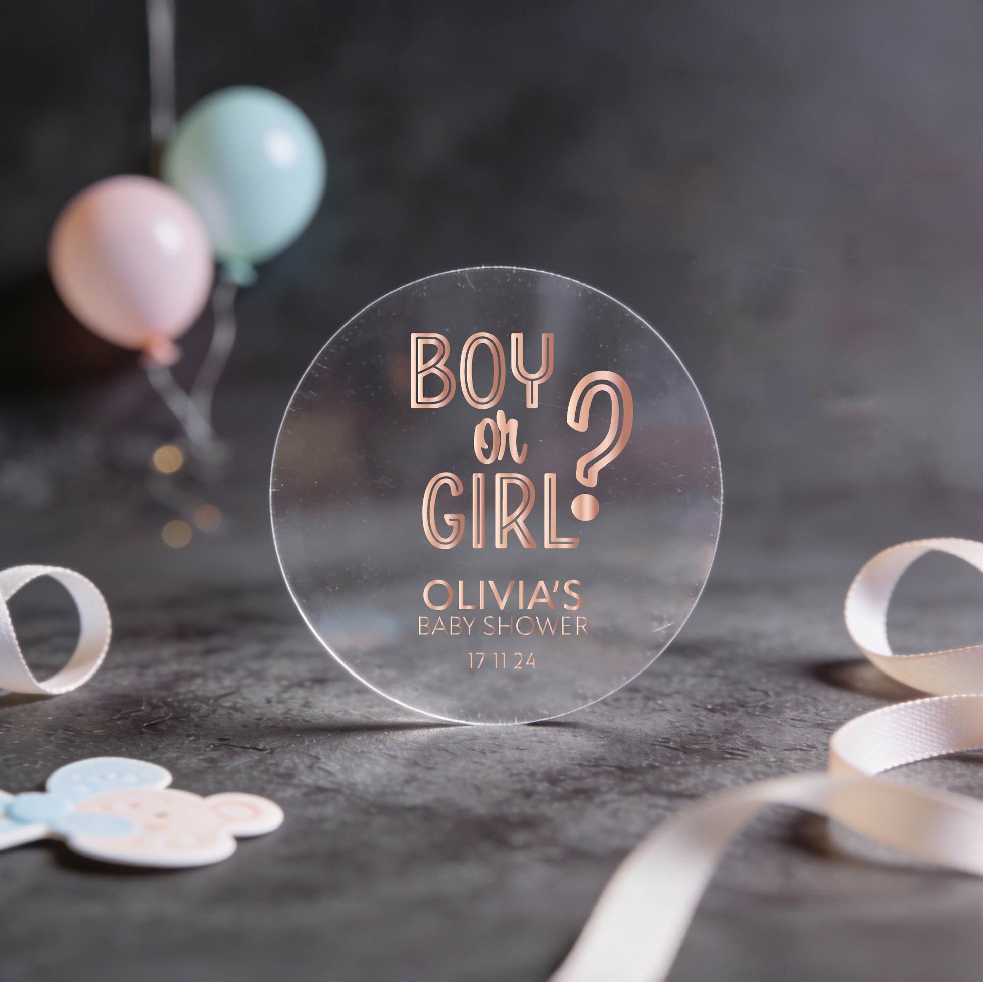 Transparent round sticker with 'Boy or Girl?' question and baby shower details written in rose gold foil.