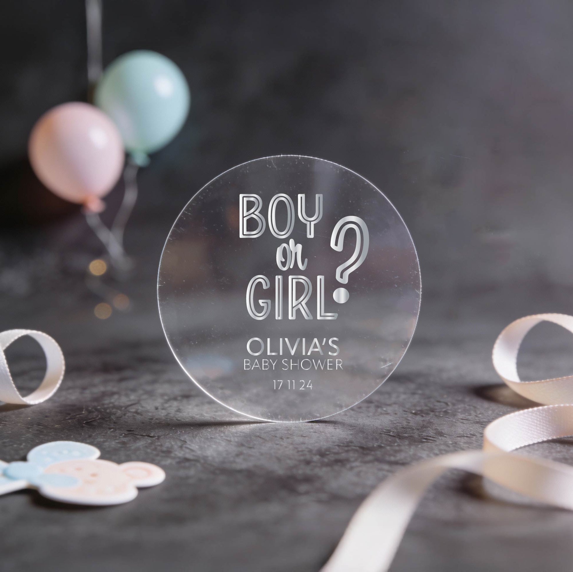 Transparent round sticker with 'Boy or Girl?' question and baby shower details written in silver foil. 