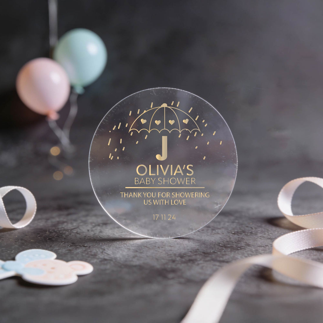 Transparent round sticker with gold foil text, “Olivia's Baby Shower,” featuring an umbrella with hearts.