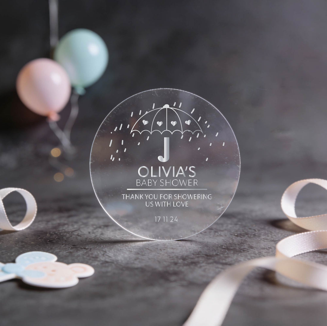 Transparent round sticker with gold foil text, “Olivia's Baby Shower,” featuring an umbrella with hearts.