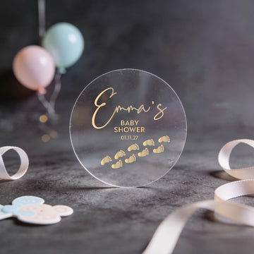 Transparent baby shower sticker with gold foil script and tiny footprints, set against a soft focus background with pastel balloons. Celebratory and elegant.