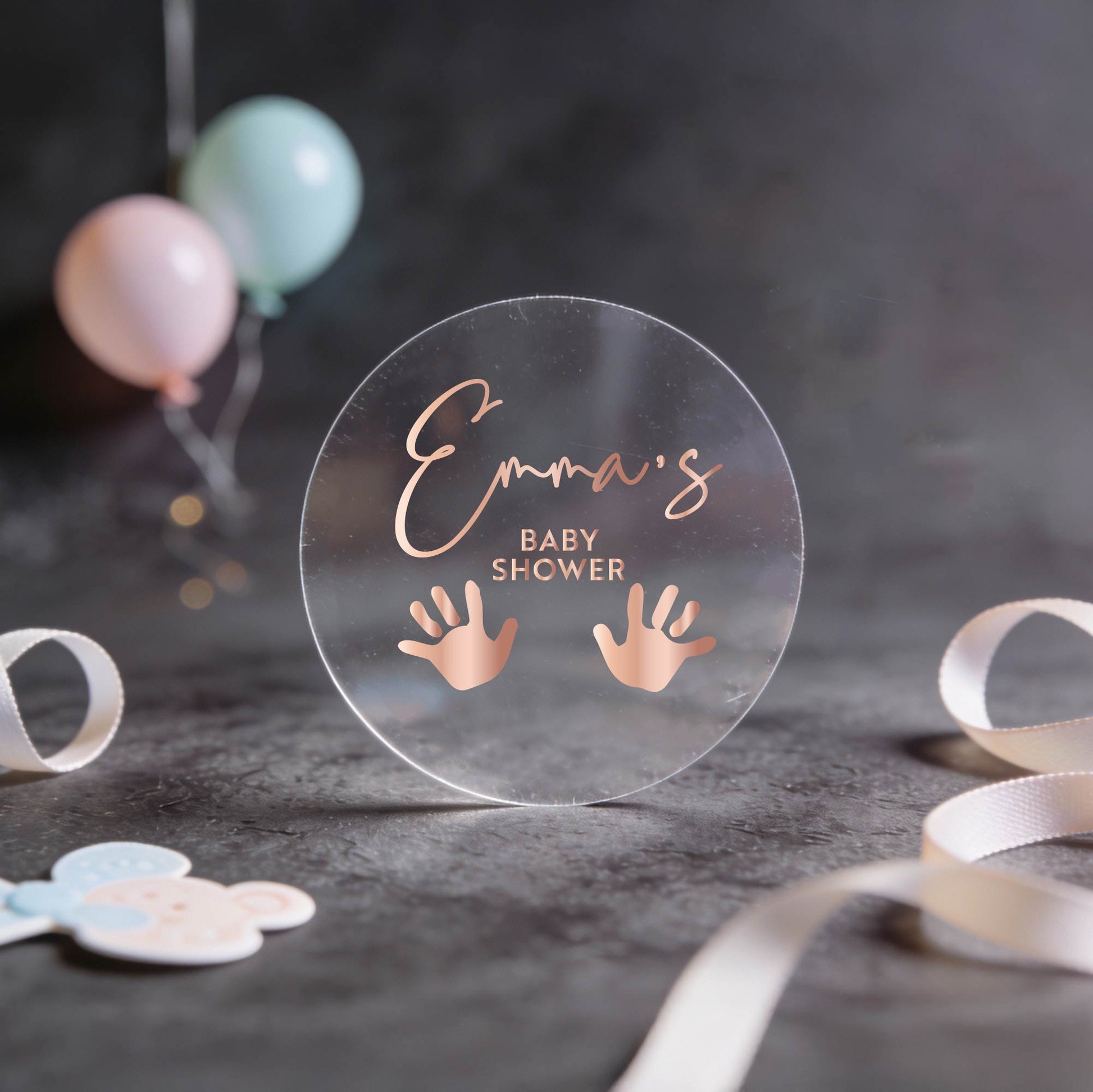 Transparent round sticker with "Emma's Baby Shower" in rose gold foil script and handprint icons. 
