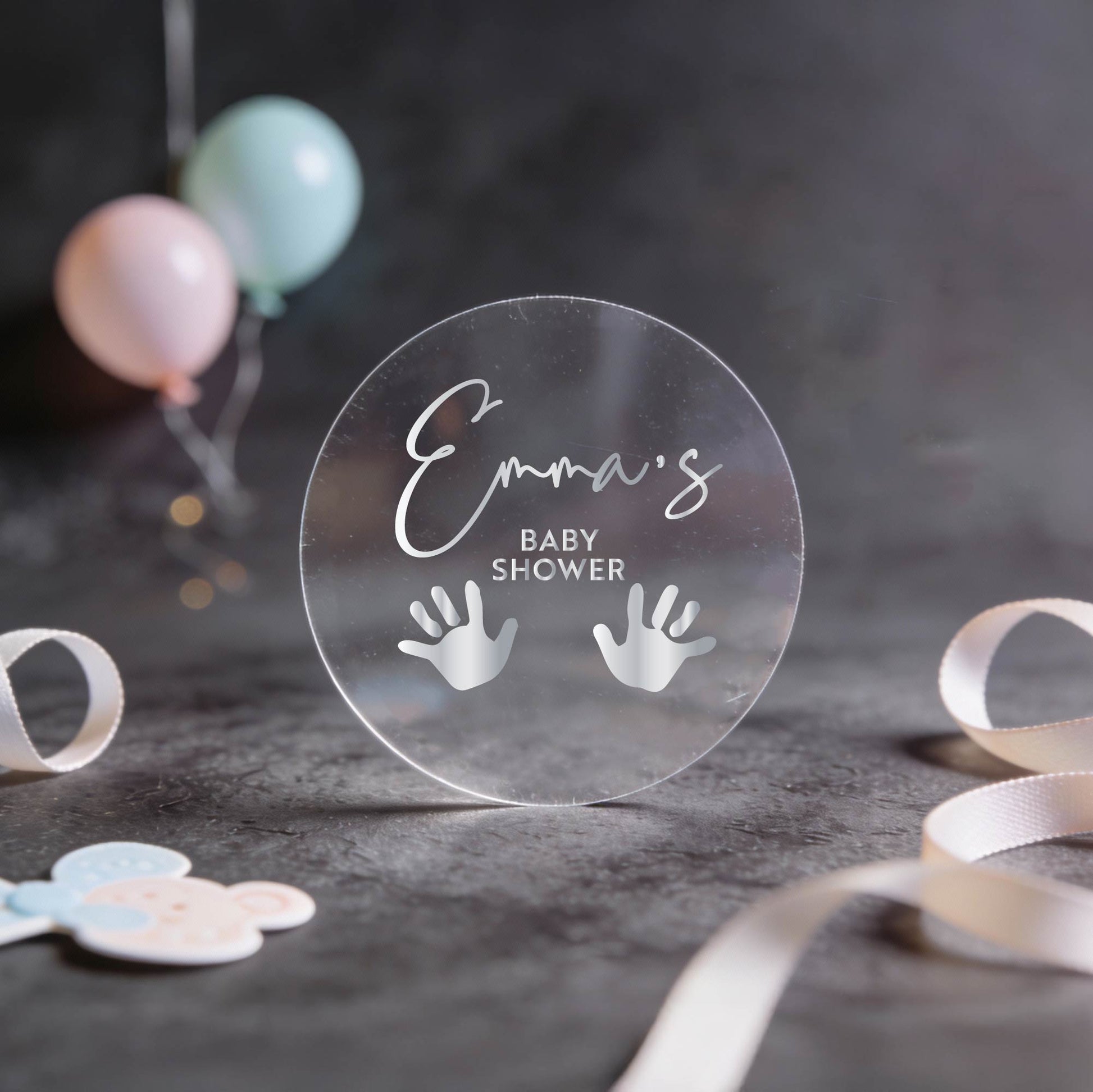 Transparent round sticker with "Emma's Baby Shower" in silver foil script and handprint icons. 