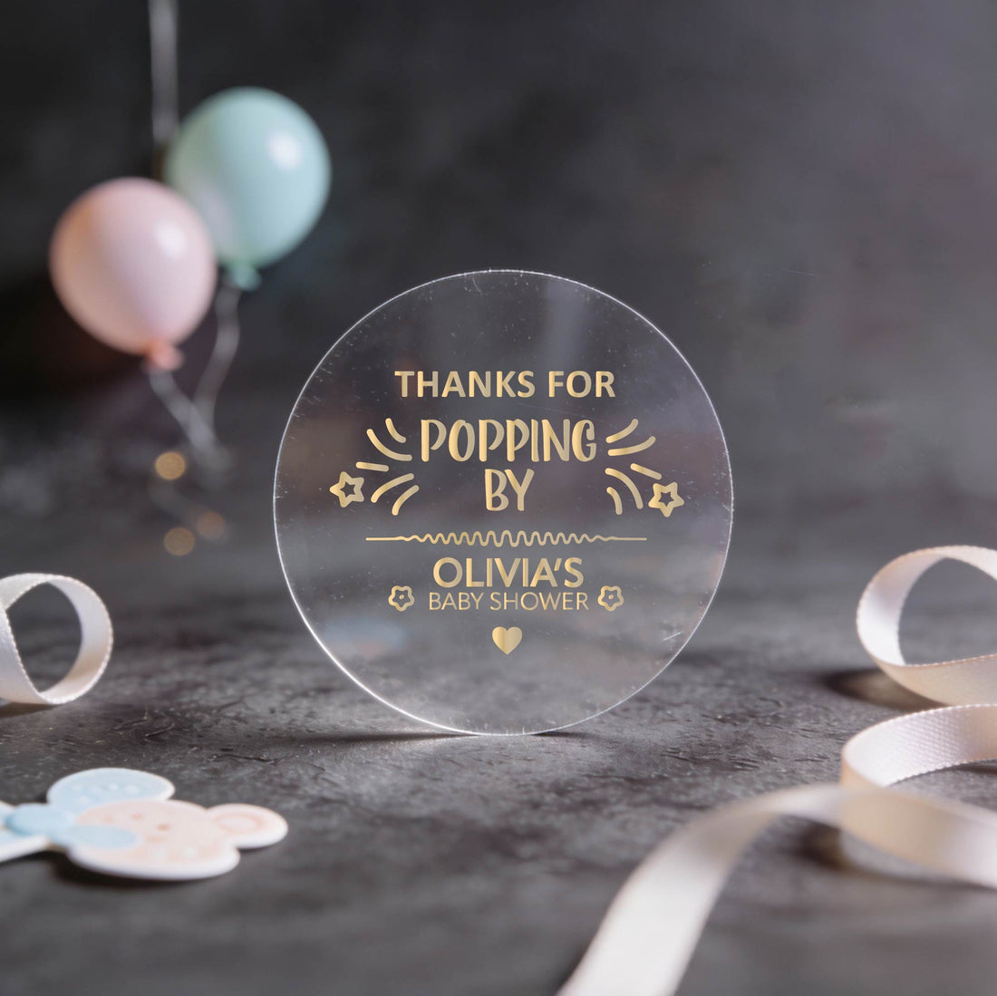 Transparent round sticker with gold foil text 'Thanks for popping by Olivia's Baby Shower' on a dark surface with balloons and ribbons.
