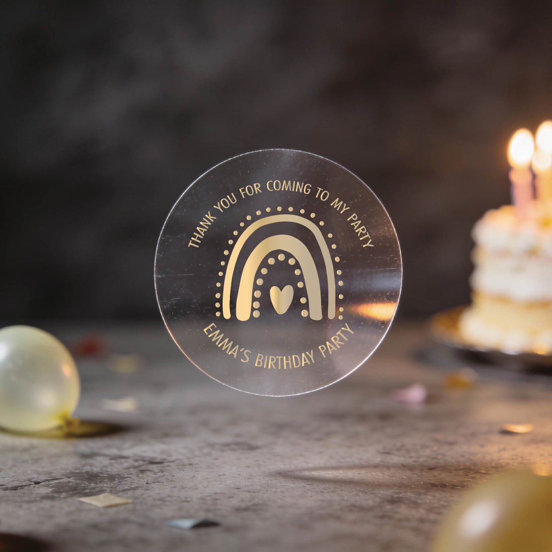 A clear round party favour sticker with a gold foil rainbow and heart design reads "Thank you for coming to my party. Emma's Birthday Party." A lit cake and balloons are blurred in the background. The setting is festive and warm.
