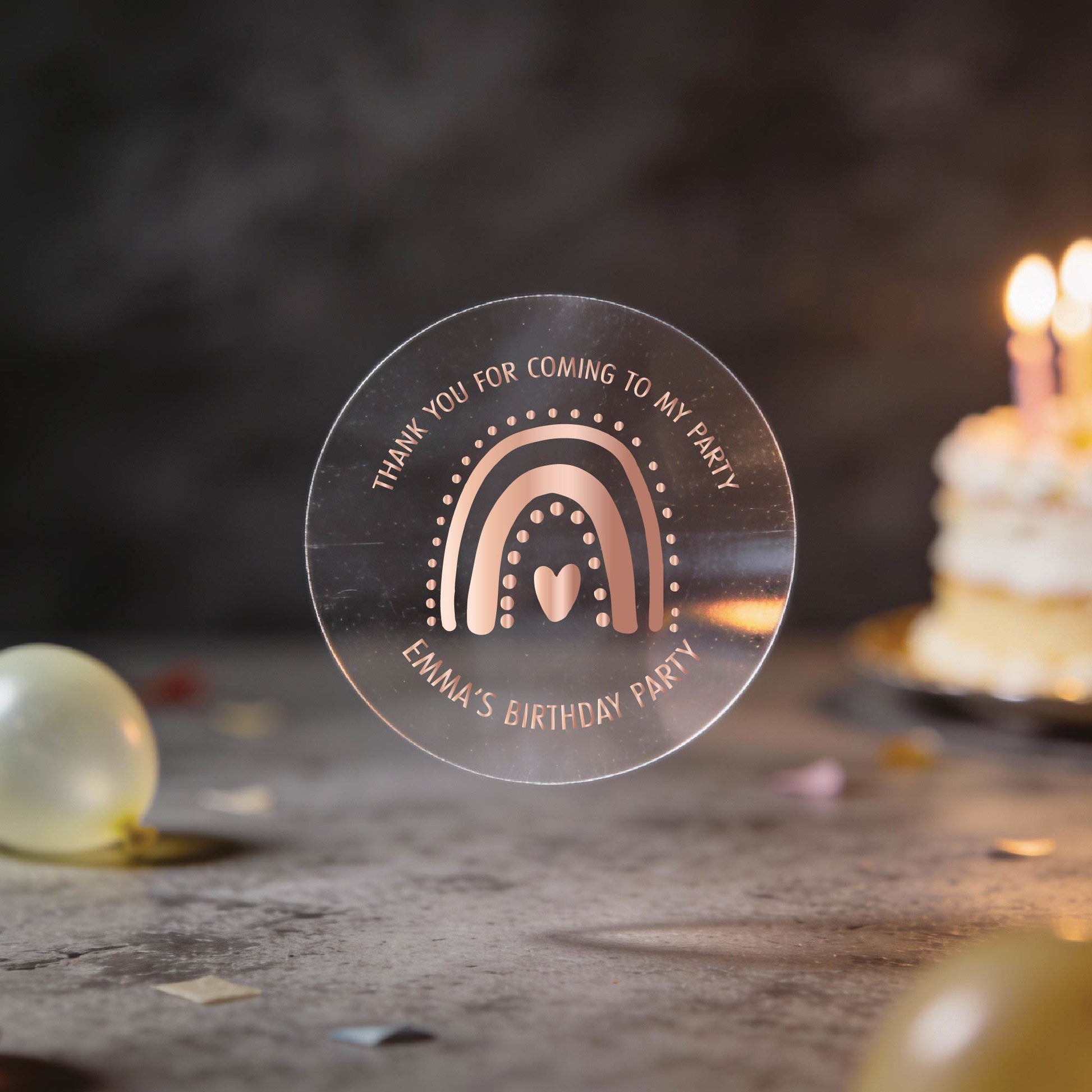 A clear round party favour sticker with a rose gold foil rainbow and heart design reads "Thank you for coming to my party. Emma's Birthday Party." A lit cake and balloons are blurred in the background. The setting is festive and warm.