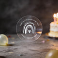 A clear round party favour sticker with a silver foil rainbow and heart design reads 