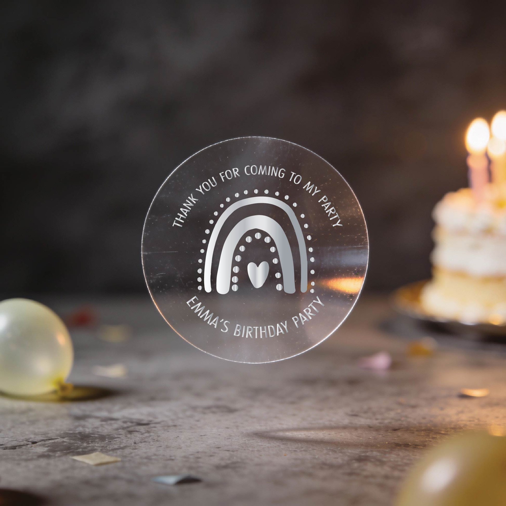 A clear round party favour sticker with a silver foil rainbow and heart design reads "Thank you for coming to my party. Emma's Birthday Party." A lit cake and balloons are blurred in the background. The setting is festive and warm.