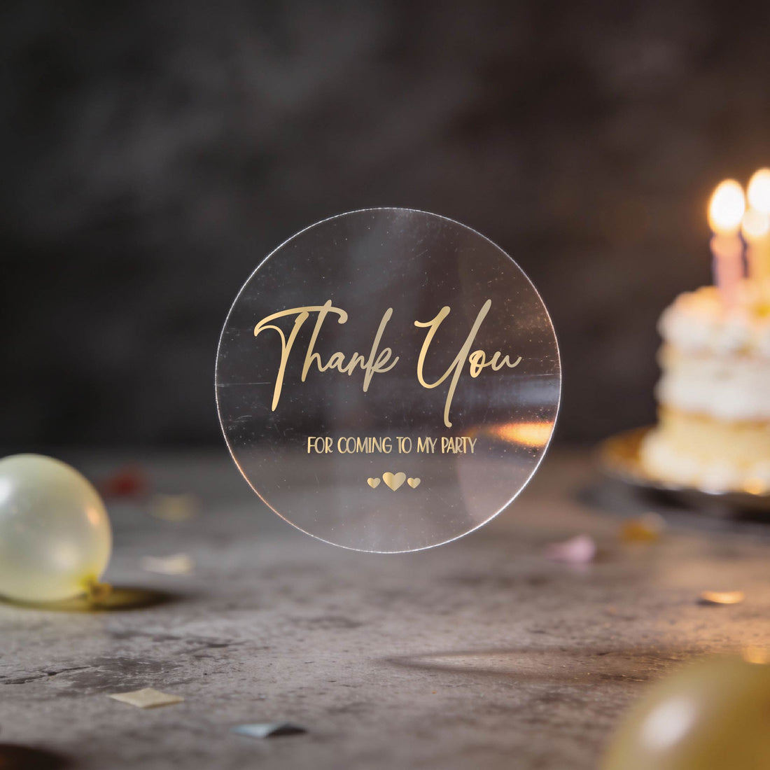 A transparent sticker with gold foil text saying "Thank You for coming to my party," against a blurred background of colourful balloons and a lit birthday cake, conveying gratitude and celebration.