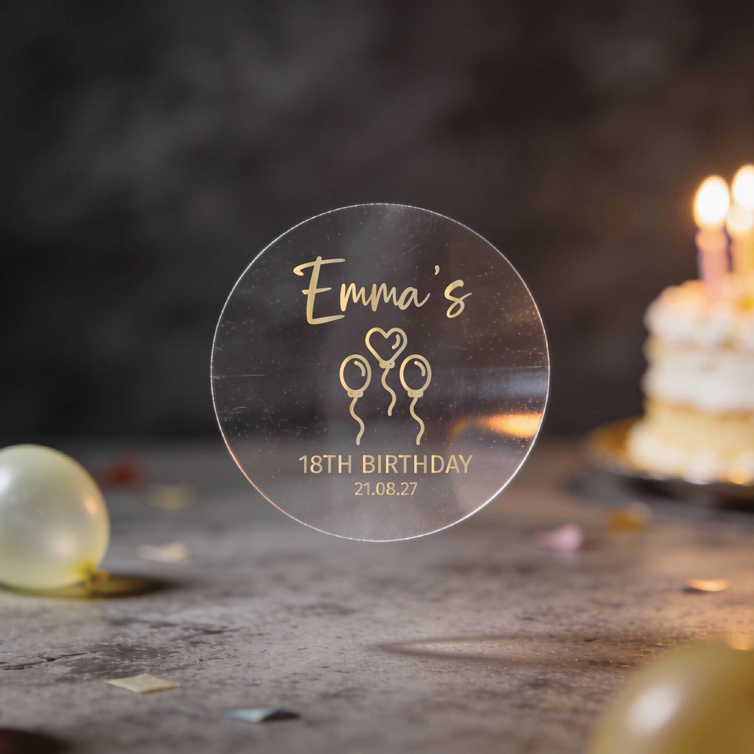 Transparent Clear sticker with gold foil text "Emma's 18th Birthday 21.08.27" and balloon icons. A lit birthday cake softly glows in the blurred background.