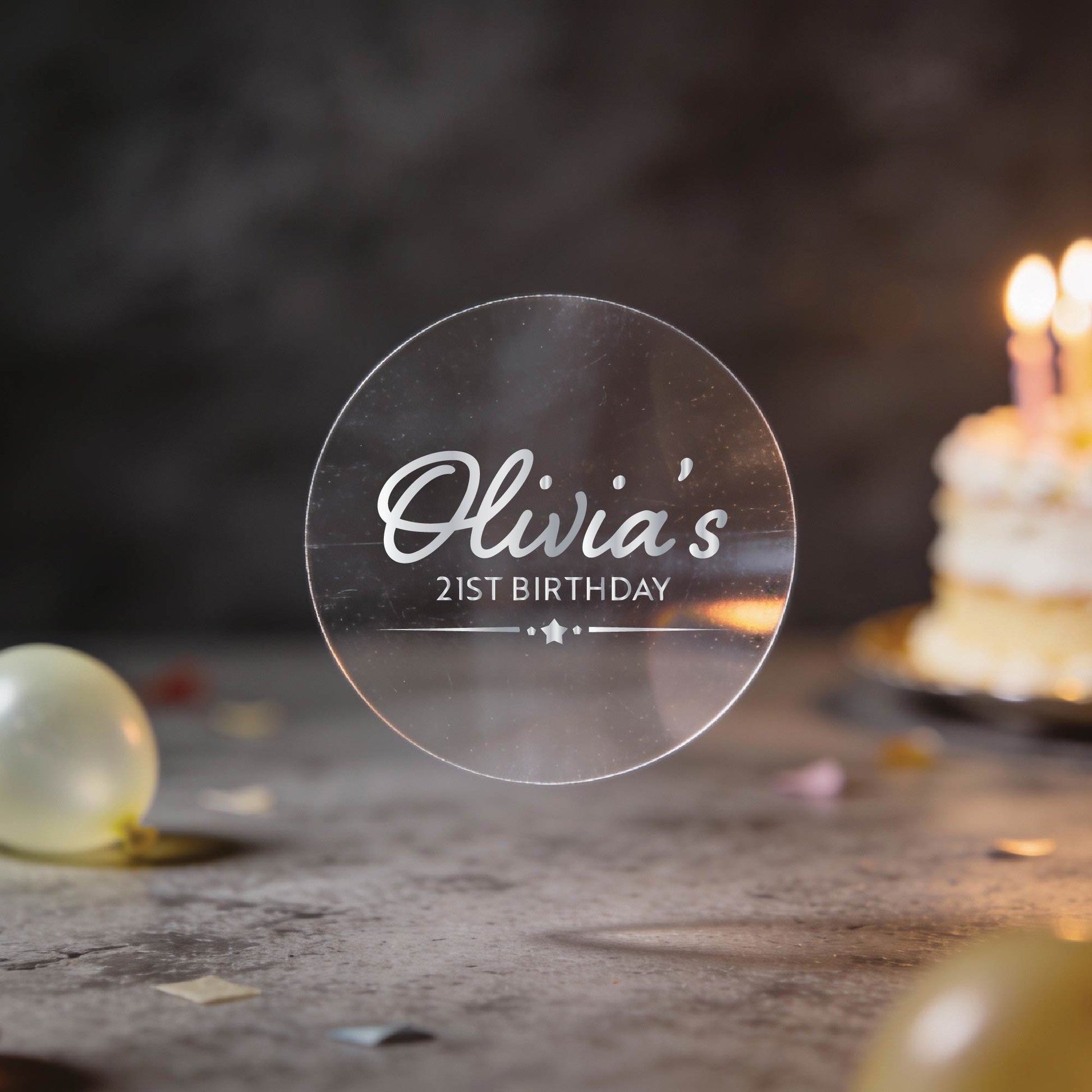 A transparent round sticker with "Olivia's 21st Birthday" in silver foil lettering with a star border line. 