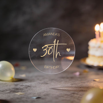 A transparent round sticker with "Amanda's 30th Birthday" in elegant script with gold foil print. The "30th" is large in the centre with two hearts either side of it.