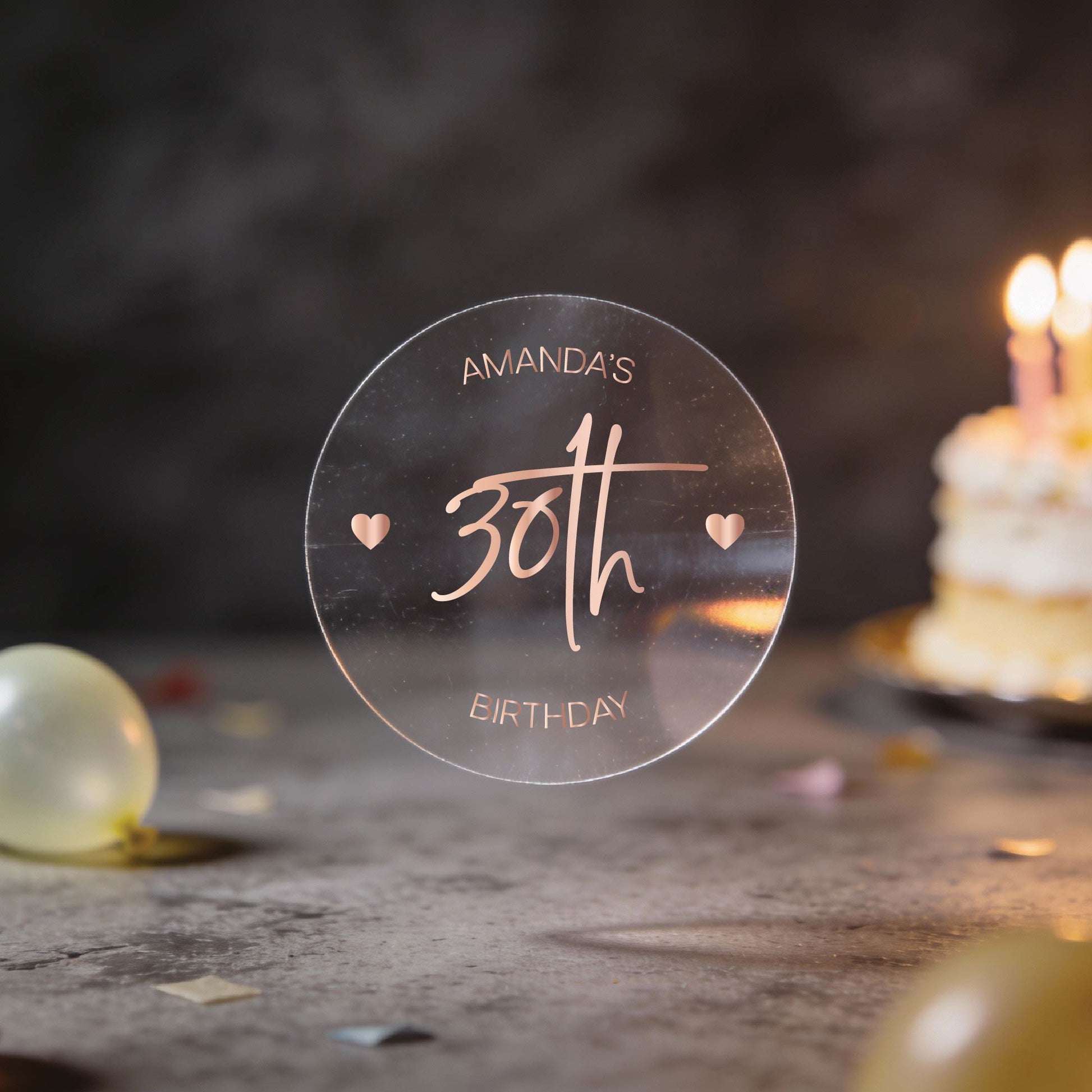 A transparent round sticker with "Amanda's 30th Birthday" in elegant script with rose gold foil print. The "30th" is large in the centre with two hearts either side of it.