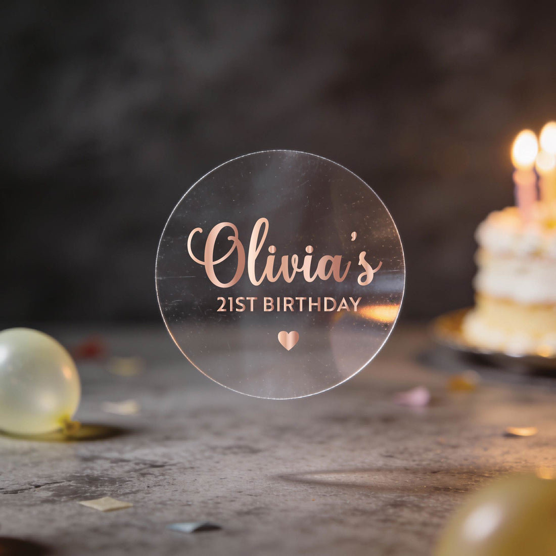 Round Transparent clear sticker with a name written in gold foil using a elegant handwritten font and milestone age under the name and heart icon to finish the sticker off. 