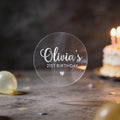 Round Transparent clear sticker with a name written in silver foil using a elegant handwritten font and milestone age under the name and heart icon to finish the sticker off. 