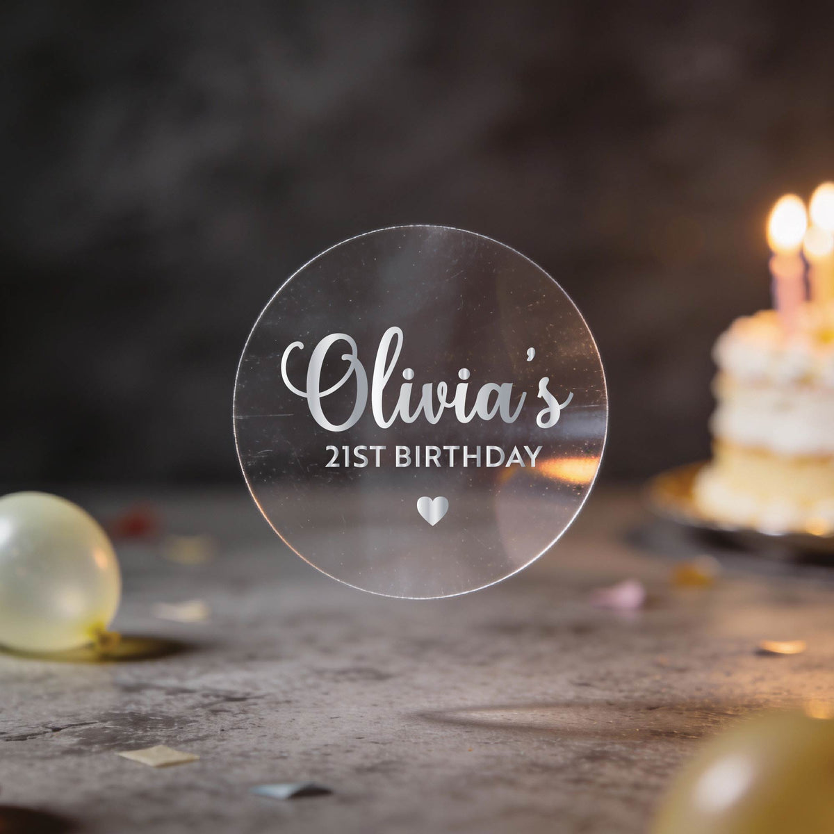 Round Transparent clear sticker with a name written in silver foil using a elegant handwritten font and milestone age under the name and heart icon to finish the sticker off. 