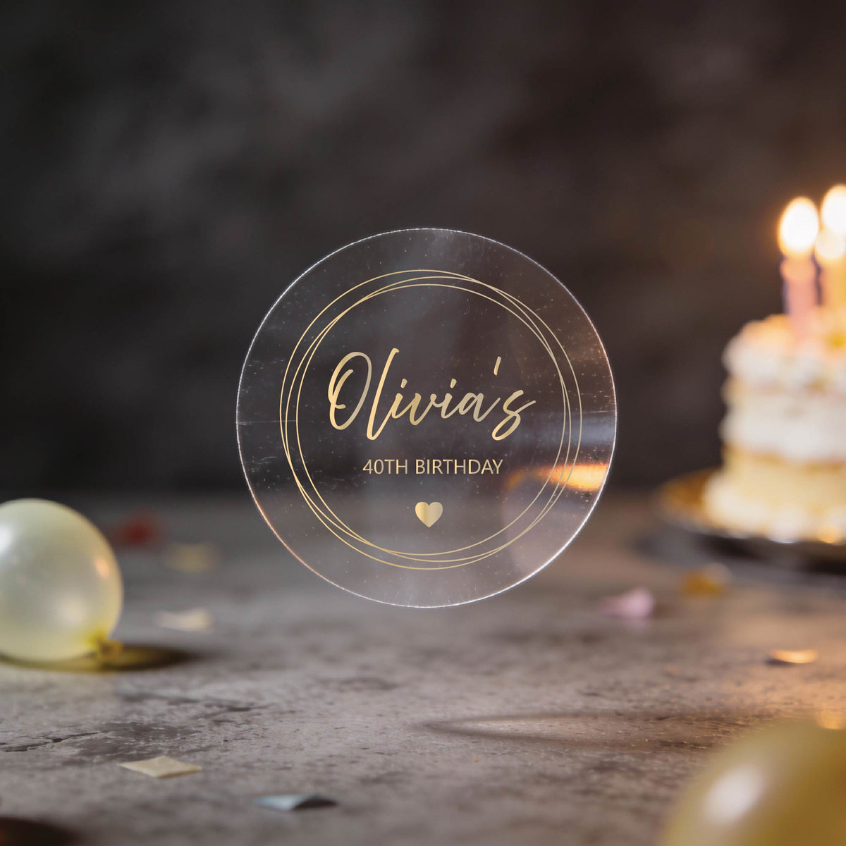 A clear round sticker with gold foil lettering that reads 'Olivia's 40th Birthday' and a small heart with a gold round frame border. 