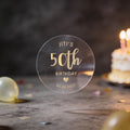 A clear round sticker with gold foil text reading 'JITI'S 50th BIRTHDAY'. A heart icon and date finishes the sticker off.