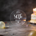 A hand holds a clear round sticker with silver foil text reading 'JITI'S 50th BIRTHDAY'. A heart icon and date finishes the sticker off.