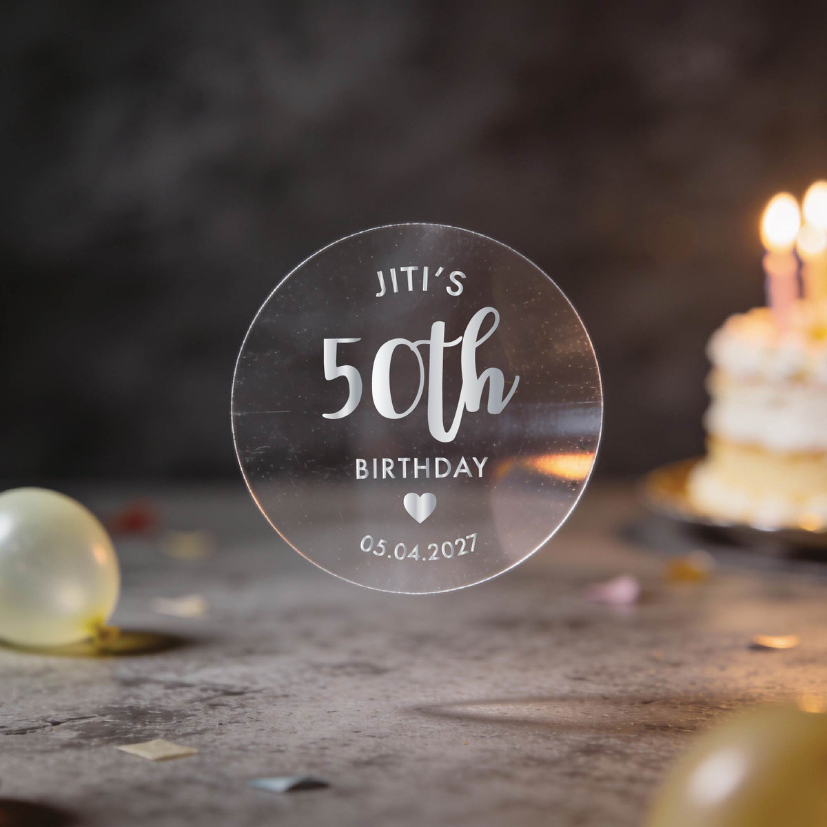 A hand holds a clear round sticker with silver foil text reading 'JITI'S 50th BIRTHDAY'. A heart icon and date finishes the sticker off.