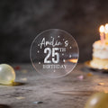 A hand holds a round transparent sticker with silver foil text reading 'Amelia's 25th Birthday' and silver stars surrounding the design
