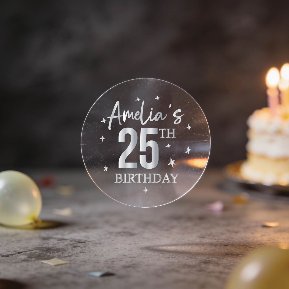 A hand holds a round transparent sticker with silver foil text reading 'Amelia's 25th Birthday' and silver stars surrounding the design