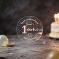 A hand holds a clear round sticker with rose gold foil text reading 'Thank you for coming to Sarah's 1derful Birthday party' against a blurred birthday scene with balloons and a lit cake.