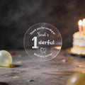 A hand holds a clear round sticker with silver foil text reading 'Thank you for coming to Sarah's 1derful Birthday party' against a blurred birthday scene with balloons and a lit cake.