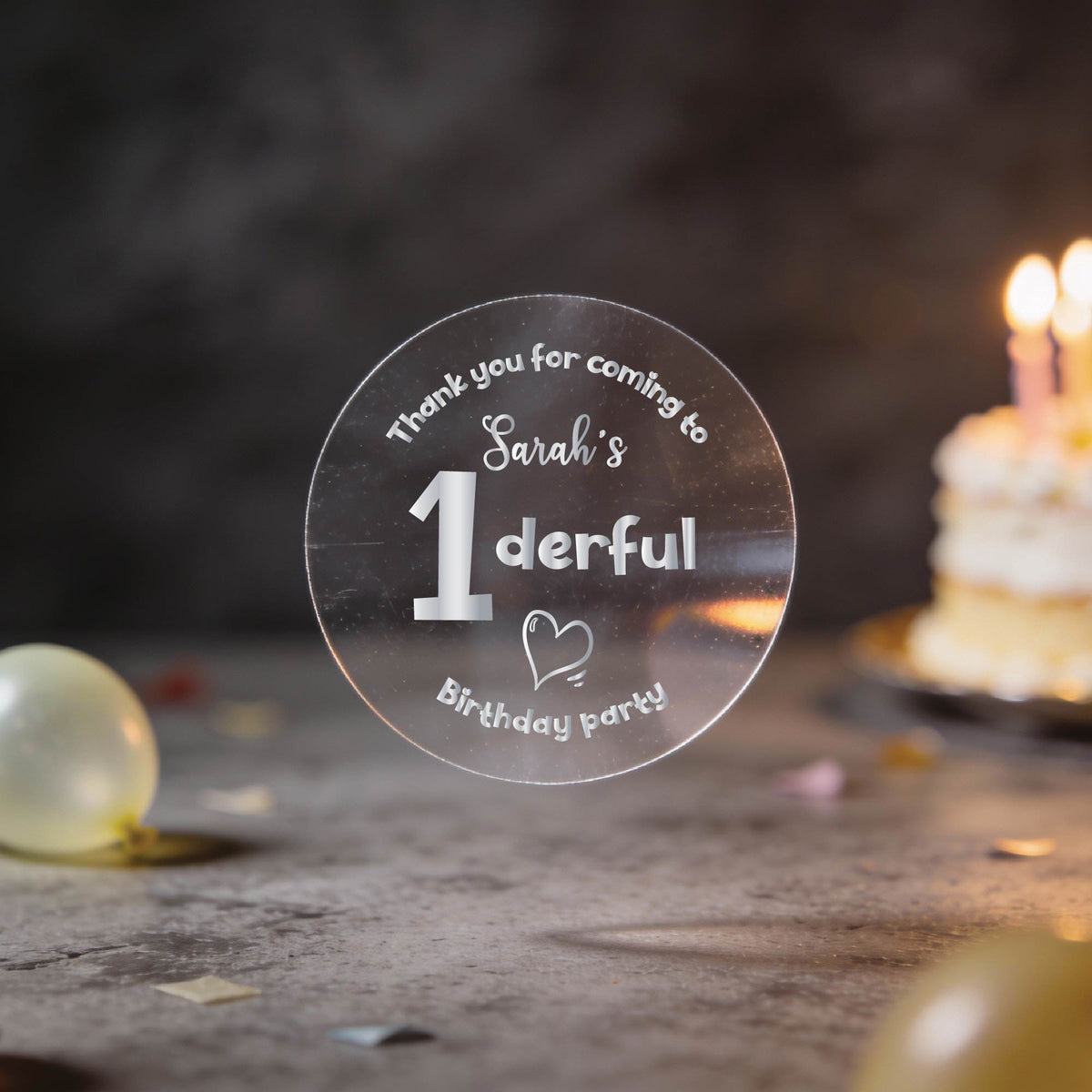 A hand holds a clear round sticker with silver foil text reading 'Thank you for coming to Sarah's 1derful Birthday party' against a blurred birthday scene with balloons and a lit cake.