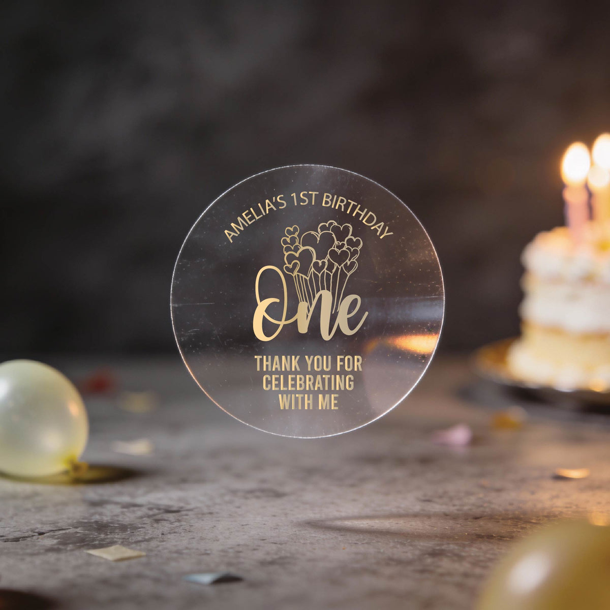 A clear round sticker with gold foil text reading 'Amelia's 1st Birthday One Thank You for Celebrating with Me', a design with the word "one" and heart balloons coming from the word. 