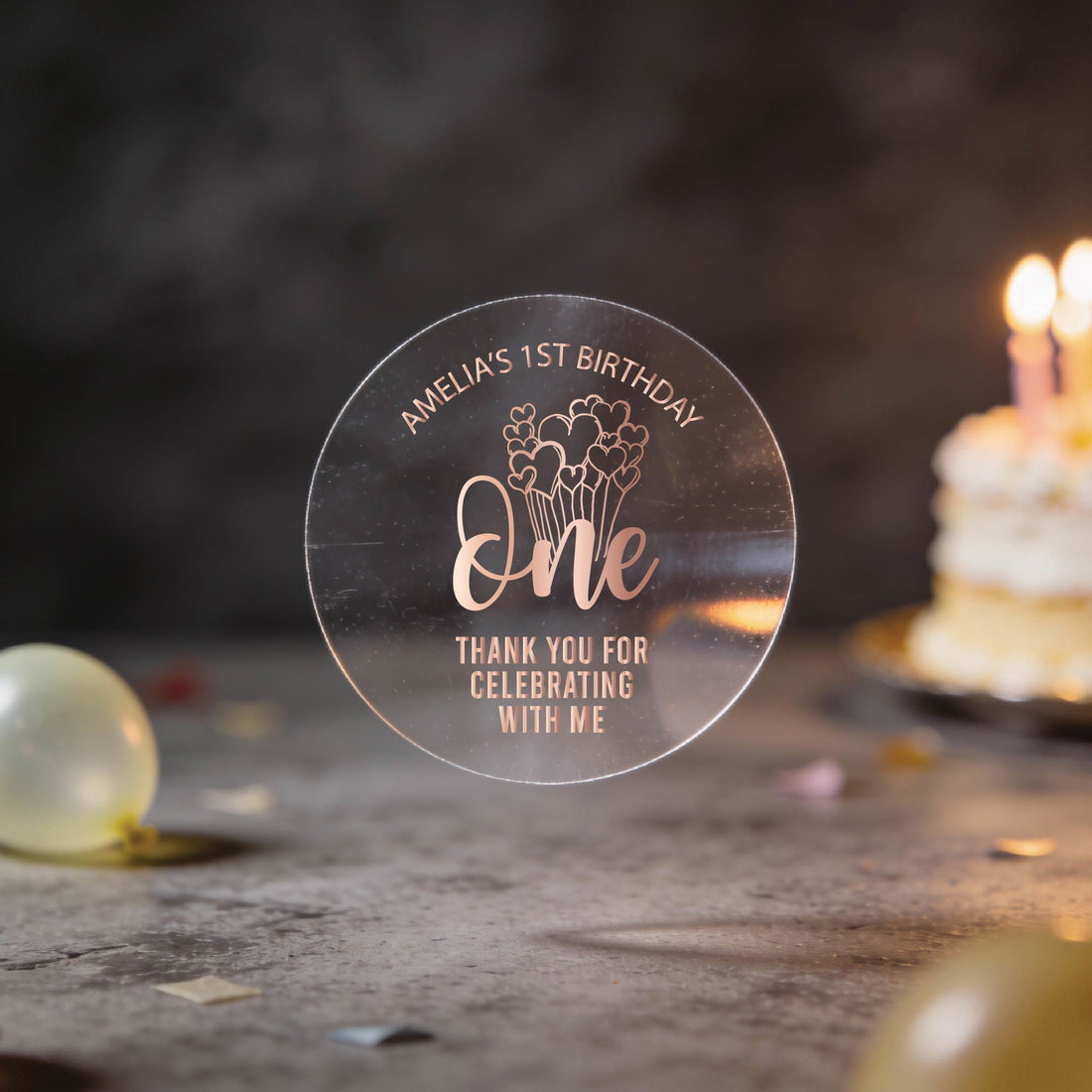 A hand holds a clear round sticker with gold foil text reading 'Amelia's 1st Birthday One Thank You for Celebrating with Me', a design with the word "one" and heart balloons coming from the word. 