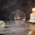 A clear round sticker with rose gold foil text reading 'Amelia's 1st Birthday One Thank You for Celebrating with Me', a design with the word 