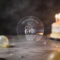 A clear round sticker with silver foil text reading 'Amelia's 1st Birthday One Thank You for Celebrating with Me', a design with the word 