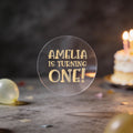 A round clear sticker with gold foil text reading 'AMELIA IS TURNING ONE!' against a dark birthday background with balloons and a blurred birthday cake.