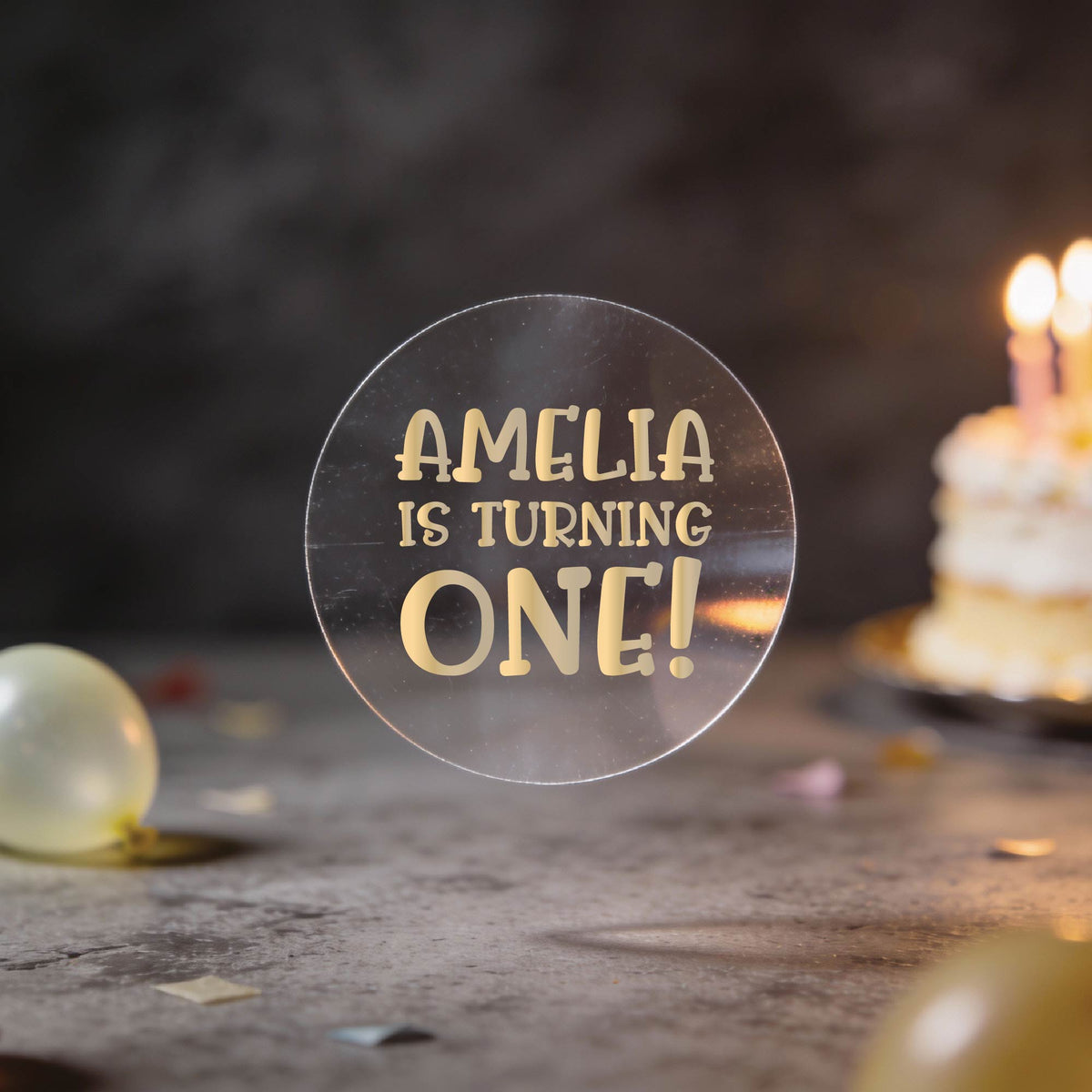 A round clear sticker with gold foil text reading 'AMELIA IS TURNING ONE!' against a dark birthday background with balloons and a blurred birthday cake.