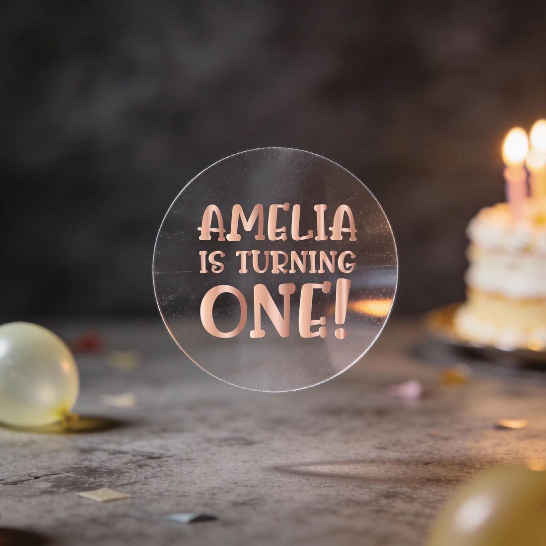 A hand holds a round clear sticker with gold foil text reading 'AMELIA IS TURNING ONE!' against a dark birthday background with balloons and a blurred birthday cake.