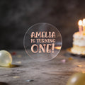 A round clear sticker with rose gold foil text reading 'AMELIA IS TURNING ONE!' against a dark birthday background with balloons and a blurred birthday cake.