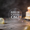 A round clear sticker with silver foil text reading 'AMELIA IS TURNING ONE!' against a dark birthday background with balloons and a blurred birthday cake.