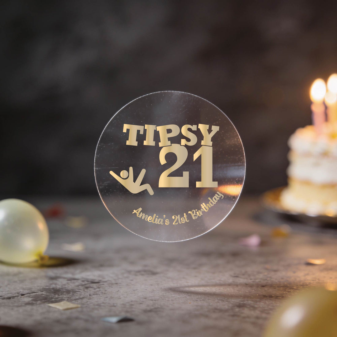 A hand holds a clear round transparent sticker with gold foil text reading 'TIPSY 21' and 'Amelia's 21st Birthday' against a dark tabletop with blurred balloons and a birthday cake in the background.