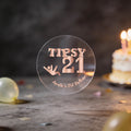 A clear round transparent sticker with rose gold foil lettering that reads 'TIPSY 21' and 'Amelia's 21st Birthday,' shown on a tabletop with a blurred birthday cake and candles in the background.