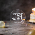 A clear round transparent sticker with silver foil lettering that reads 'TIPSY 21' and 'Amelia's 21st Birthday,' shown on a tabletop with a blurred birthday cake and candles in the background.