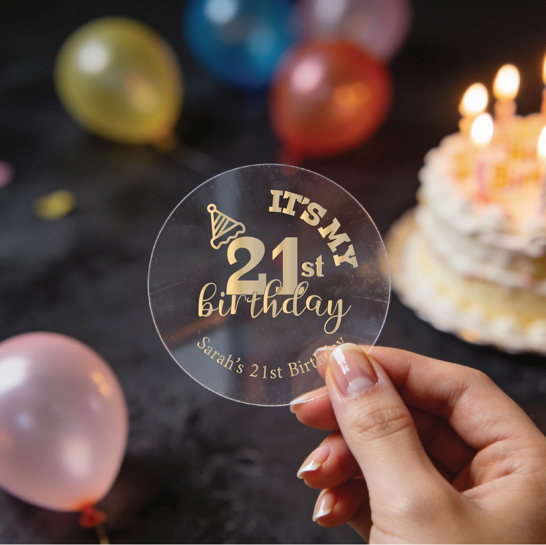 A hand holds a clear round with gold foil lettering that reads 'It's my 21st Birthday' & 'Sarah's 21st Birthday'. There is a party hat icon on the number 2. 