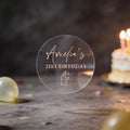 A hand holds a clear round sticker with rose gold foil text saying 'Amelia's 21st Birthday' and a little present icon underneath. 