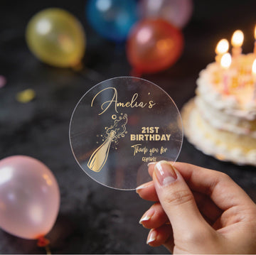 A hand holds a round clear sticker with gold foiling saying 'Amelia's 21st Birthday Thank you for coming' and a champagne bottle illustration, with blurred birthday balloons and a lit cake in the background.